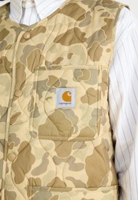Kviltad väst i tan kamouflage-mönster, med en framficka som har ett Carhartt-logga. Lätt tyg med slät textur och knäppning.