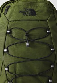 Sac à dos en tissu vert avec un logo noir, des cordons élastiques et des éléments métalliques. Surface texturée avec des coutures durables.
