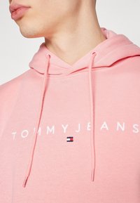 Světle růžová mikina s kapucí s předním logem a bílým textem "TOMMYJEANS" a malou barevnou nášivkou. Šňůrky dodávají designu šmrnc.
