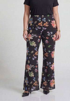 Kvinde iført sorte bukser med vide ben og farverigt blomsterprint, kombineret med sorte sko og en sort kortærmet top.