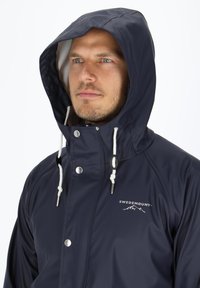 Navy Regenjacke mit Kapuze, ausgestattet mit silbernen Druckknöpfen und weißen Kordeln. Das Logo "SWEDEMOUNT" ist auf der Brust aufgedruckt. Glatte Textur.