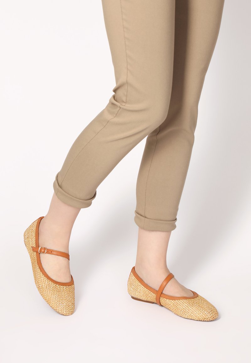 Tan ballerina flats van geweven stroachtig materiaal, met een ronde neus en een bruine enkelband. De zolen zijn plat voor comfort.