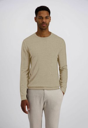 Mann steht mit den Händen in den Taschen, trägt einen beigen Langarmpullover und helle beiger Hose vor einem schlichten Hintergrund.
