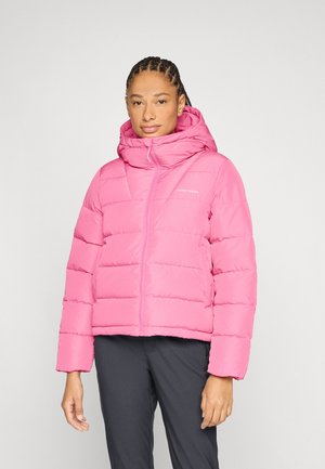 Femme debout portant une doudoune rose vif avec capuche et un pantalon noir, sur un fond clair uni.