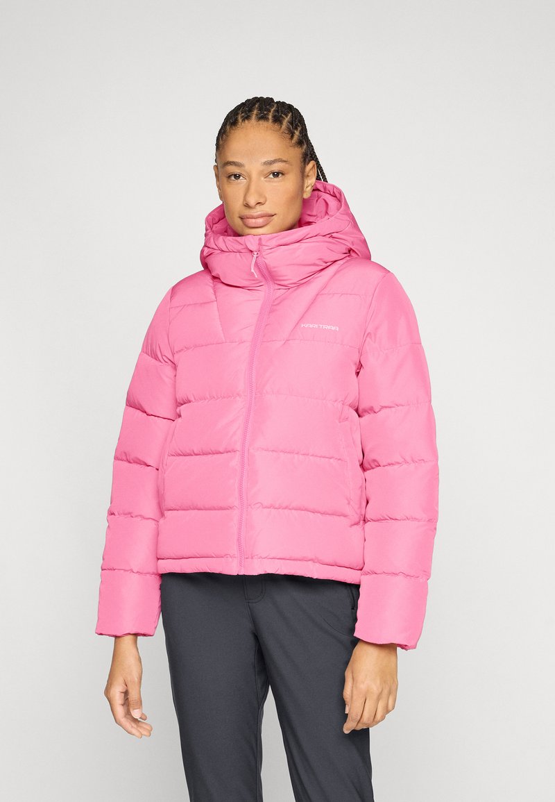 Femme debout portant une doudoune rose vif avec capuche et un pantalon noir, sur un fond clair uni.