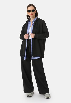 Black hooded sweatshirt, layered over a light blue striped shirt, paired with black wide-leg pants and gray sneakers. Accessorized with sunglasses.