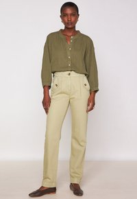 Vrouw staat gekleed in een olijfgroene gestreepte blouse met knopen, beige hoge taille broek met knoopzakken en donkerbruine platte schoenen.