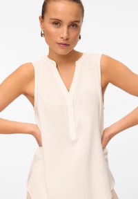 Blusa senza maniche di colore crema con colletto alto, chiusura a bottoni e silhouette fluttuante con spacchi laterali per una maggiore libertà di movimento.