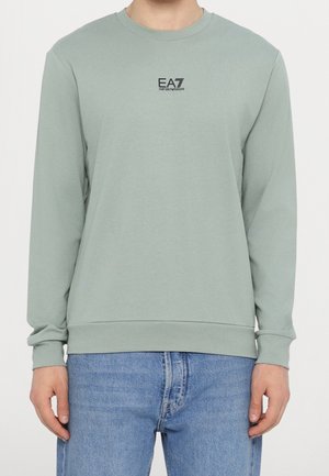 Mężczyzna w jasnozielonej bluzie z długim rękawem z logo EA7 Emporio Armani na piersi i jasno-niebieskich dżinsach, stojący z rozluźnionymi ramionami.