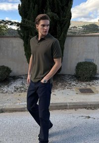 Mladý muž v tmavozelenom polu a džínsoch stojí na chodníku, ruky v vreckách, na pozadí vysokých stromov a kamennej steny.