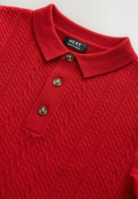 Röd stickad polo tröja med ett strukturerat diamantsmönster, tre bruna knappar och en platt krage. Etiketten visar "NEXT, MADE IN MYANMAR."