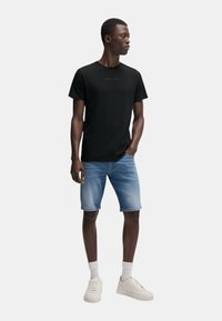 T-shirt nera di Calvin Klein, pantaloni corti di jeans chiari e sneakers bianche con lacci visibili. Il modello è in piedi contro uno sfondo neutro.