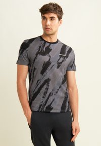 Hombre vestido con una camiseta de manga corta de patrones abstractos en negro y gris, y pantalones negros, de pie con las manos en los bolsillos frente a un fondo claro.