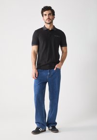 Schwarzes Polohemd mit kurzen Ärmeln und grauem Besatz, kombiniert mit lässigen blauen Jeans und schwarzen Schuhen mit weißen Streifen.