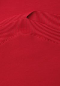 Close-up van de halslijn van een rode gebreide trui met een geribde kraag en gestikte naaddetails op gladde stof.