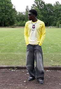 Dzeltens garām piedurknēm Nike krekls ar strukturētu audumu un logo, kombinēts ar vaļīgām, izbalējušām melnām džinsām un melnām kedām, stāv uz zāles.