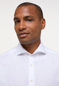 Camicia di lino bianca con colletto classico e cinque bottoni. Il tessuto ha un aspetto leggero e strutturato, adatto per il clima caldo.