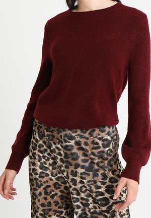 Femme portant un pull bordeaux à manches longues et un pantalon imprimé léopard sur fond blanc uni.