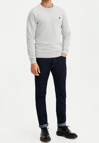 Ljusgrå texturerad tröja med rund halsringning och liten gul logotyp, kombinerad med mörkblå denimjeans och svarta finskor.