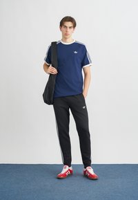 Námorníckomodrá tričko s bielymi pruhmi na rukávoch, čierne teplákové nohavice s bielymi bočnými pruhmi a červené športové topánky. Čierna taška cez rameno.