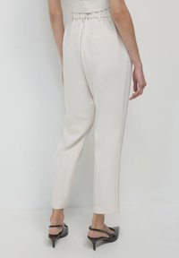Pantaloni alta vita di colore crema, con cinturino elastico, design a gamba affusolata e due tasche posteriori. Indossati con scarpe col tacco nere.