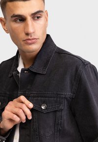 Zwarte denimjas met een klassieke snit, zichtbare stiksels, drukknopen en een borstzak. Glad textiel met een gestructureerde kraag.