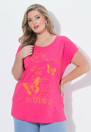 Frau mit langen blonden Haaren, die ein rosa T-Shirt mit Schmetterlingsprint und hellblaue Hosen trägt, posiert mit einer Hand in der Hüfte und der anderen nahe am Kragen.