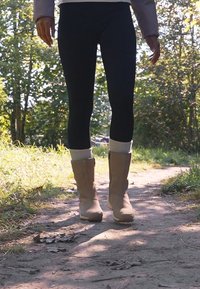 Braune Wildlederstiefel mit weißen Strickbündchen, getragen über schwarzen Leggings, auf einem Waldweg, umgeben von Grün.