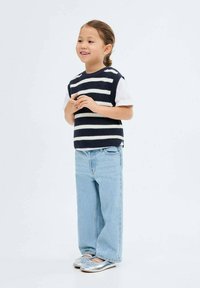 Maglione senza maniche a righe blu navy e bianche sopra una camicia bianca, abbinato a jeans azzurri e ballerine argentate. Posizione in piedi.