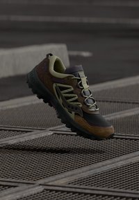 Terränglöpningsskor med en övre del i svart och olivgrönt, bruna accenter, texturerad sula och framträdande logodetaljer.