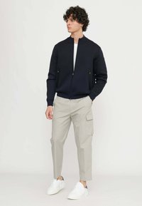 Giacca bomber blu navy con colletto alto, chiusura a zip, tasche laterali, abbinata a pantaloni cargo beige chiaro e sneaker bianche.