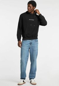 Felpa nera in cotone con coulisse e tasca frontale, abbinata a jeans azzurri e scarpe da ginnastica bianche, che mostra una vestibilità comoda e uno stile casual.