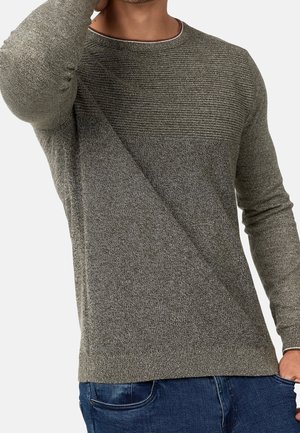 Homme portant un pull gris texturé avec côtes horizontales en haut, associé à un jean bleu, main près de la tête et dans la poche.
