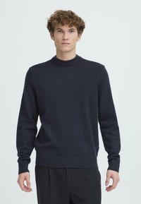 Pulover bleumarin cu guler rotund, realizat dintr-un material tricotat moale, având mâneci lungi și manșete și tiv cu nervuri, purtat peste pantaloni închiși la culoare.