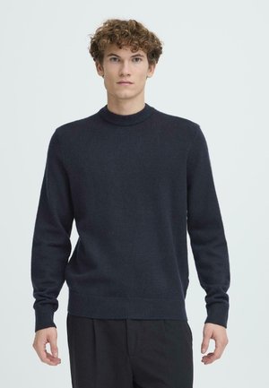 Marineblauer Rundhalsausschnitt-Pullover aus weichem Strickmaterial, mit langen Ärmeln sowie gerippten Bündchen und Saum, getragen über dunklen Hosen.