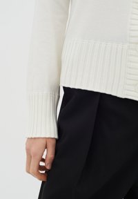 Bílý pletený kardigan s hřebenovými manžetami a lemem, s detaily knoflíků, skombinovaný s černým oděvem. Důraz na texturu a barevný kontrast.