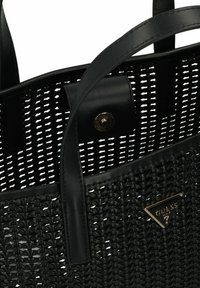 Zwarte geweven totebag met gladde leren handvatten, een magnetische drukknoopsluiting en een metalen merkembleem aan de zijkant. Gelaagd ontwerp.