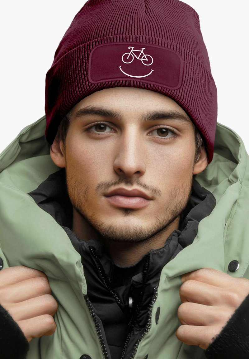 Maroon knit beanie with a smooth patch featuring a white bicycle and smile graphic. Worn with a green puffer jacket.
