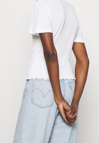 Top corto blanco de manga corta con cintura fruncida. Presenta un dobladillo con forma de ondas y tejido texturizado, combinado con unos jeans de mezclilla azul claros.