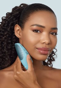 Un appareil de nettoyage facial en silicone teal avec des rainures texturées, maintenu contre la joue, présentant un design courbé et ergonomique.
