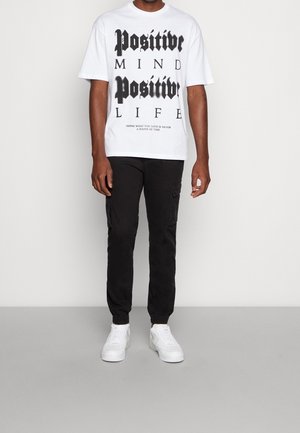 Weißes Baumwoll-T-Shirt mit schwarzem Drucktext, kombiniert mit schwarzen Cargo-Hosen und weißen Turnschuhen. Das T-Shirt verfügt über ein auffälliges Grafikdesign.