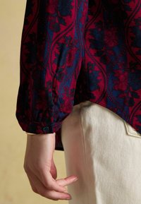 Dłoń spoczywa obok jasnobeżowych spodni, na sobie ma koszulę z długim rękawem w czerwono-niebieski kwiatowy wzór z zapięciem na guzik.