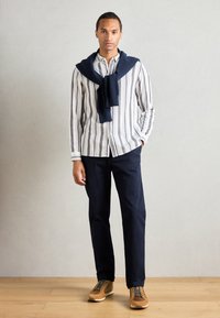 Gestreiftes Langarmhemd in Weiß und Blau, marineblauer Pullover über die Schultern gelegt, dunkle Hose, braune Schuhe mit grünen Schnürsenkeln.