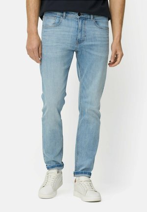 Mężczyzna w jasnoniebieskich wąskich dżinsach i białych trampkach, stojący z rękami swobodnie opuszczonymi wzdłuż ciała.