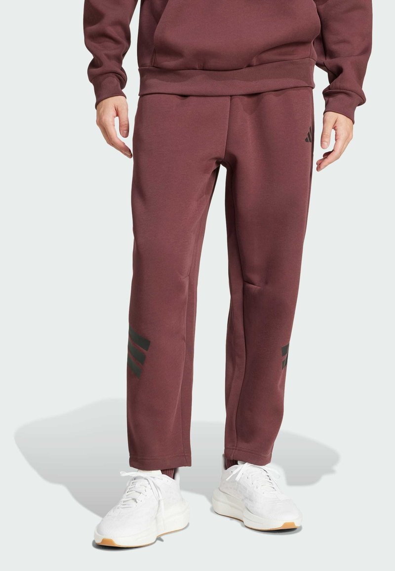 Pantalons de survêtement bordeaux en tissu doux, avec une coupe décontractée et trois rayures noires sur le bas des jambes. Associés à des chaussures de sport blanches.