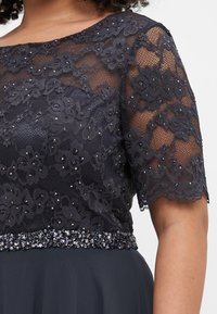 Rochie de marine cu top din dantelă cu motive florale și accente din strasuri, completată de un brâu cu mărgele. Mâneci scurte, transparente.