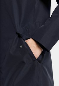 Marinblå vattentät jacka med en slät yta, med en sidoficka och knappknäppning. Logotypen "Didriksons" finns nära fickan.