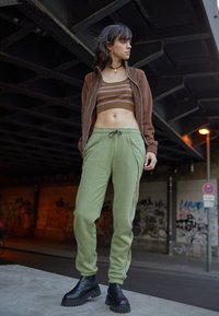 Brun zip-jacka, kort brun randig topp, gröna sweatpants med dragsko, svarta chunky stövlar. Urban miljö med graffiti som bakgrund.