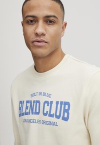 Krem sweatshirt med blå tekst "BLEND CLUB" og "LOS ANGELES ORIGINAL," laget av mykt stoff med klassisk rund hals-design.