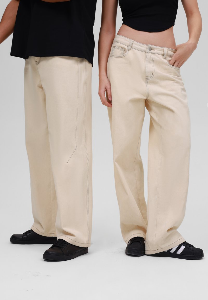 Beige wide-leg jeans with visible stitching details. Worn with black sneakers. Smooth texture, casual design, featuring standard pockets.